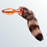 The Tailz Fox Tail Glass Anal Plug by Tailz features an orange, brown, and black striped faux furry tail attached to a transparent borosilicate glass plug, creating a whimsical animal-like appearance against a light gradient background.